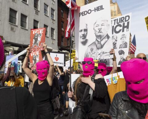 Para pengunjuk rasa yang mengenakan topeng ski berwarna merah muda memegang poster yang mengkritik Donald Trump dan ICE selama demonstrasi jalanan.