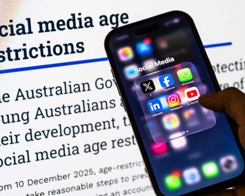 Ponsel pintar yang memegang tangan menampilkan aplikasi media sosial di depan teks tentang pembatasan usia media sosial Australia.