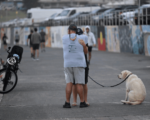 Dua orang berpelukan di jalan beraspal dengan seekor Labrador kuning duduk diikat di dekatnya dan sebuah sepeda diparkir di pagar