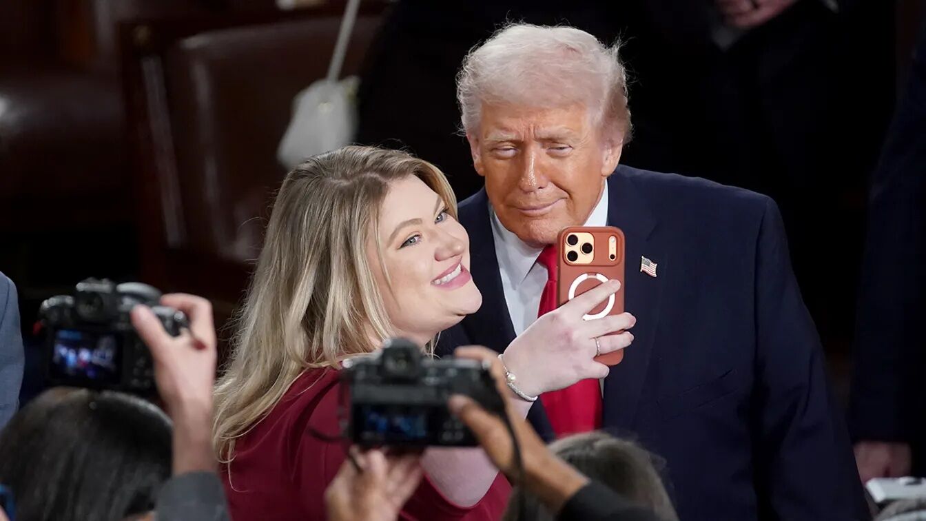Donald Trump berpose untuk selfie dengan seorang wanita tersenyum yang memegang smartphone di tengah keramaian.