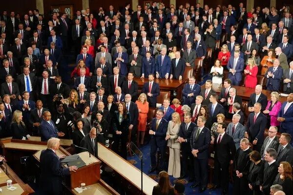 Sekelompok besar orang berpakaian formal berdiri dan mendengarkan pembicara di podium ruang legislatif.