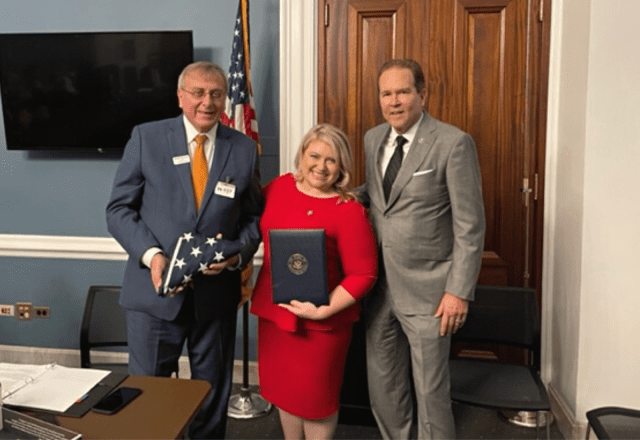 Tiga orang berpose di dalam ruangan, satu orang memegang bendera Amerika terlipat dan satu lagi memegang map sertifikat.