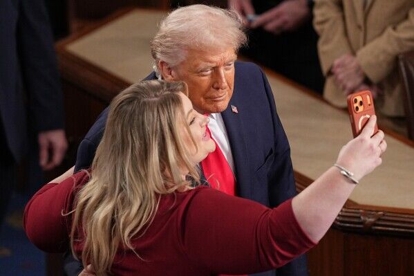 Donald Trump berpose untuk selfie dengan seorang wanita mengenakan atasan merah dalam suasana formal.