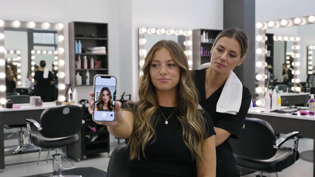 Wanita berambut panjang bergelombang berfoto selfie di salon sementara penata rambut berdiri di belakangnya dengan handuk di bahunya.