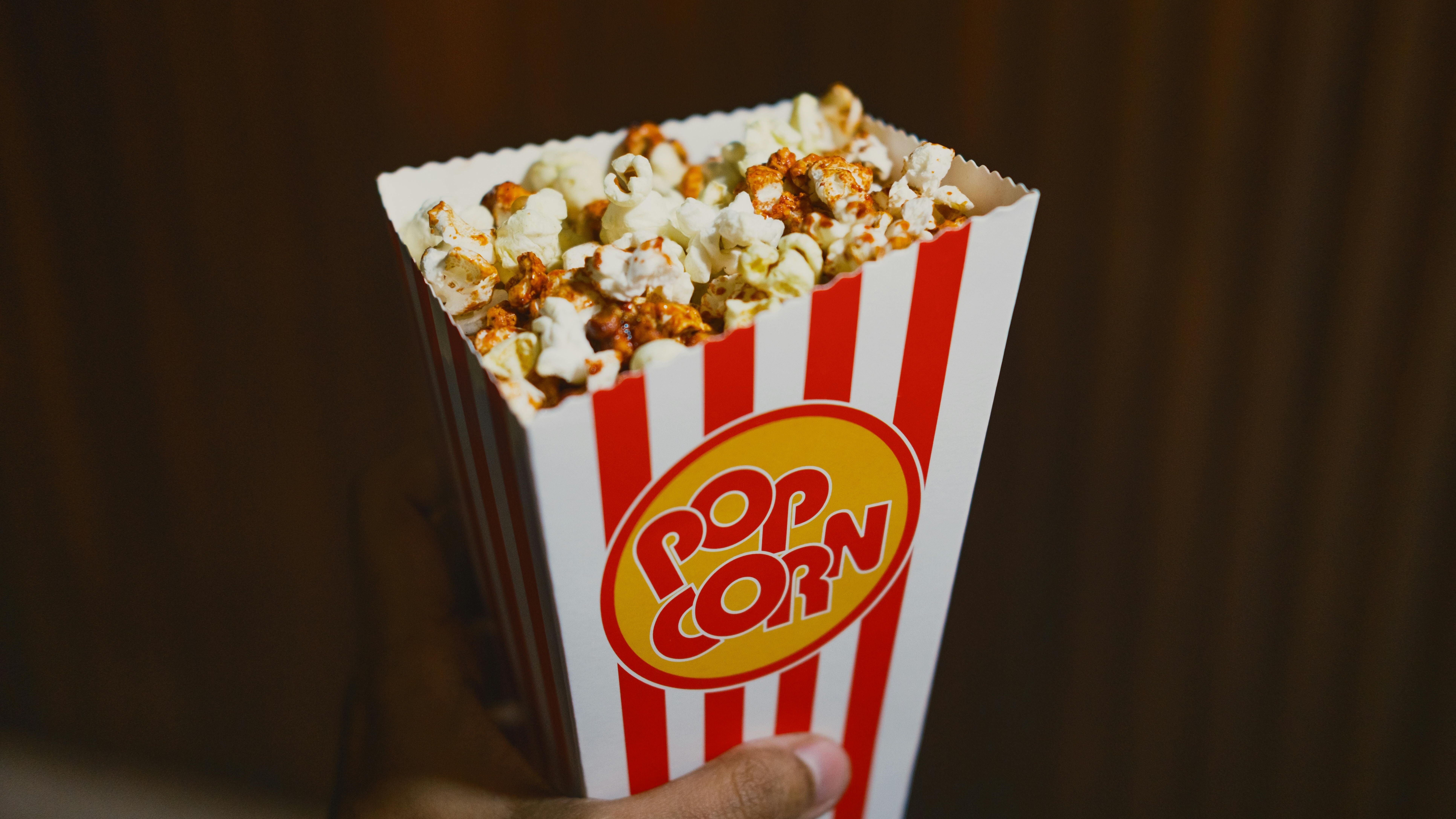 Tangan memegang kotak popcorn bergaris merah putih berisi popcorn karamel.