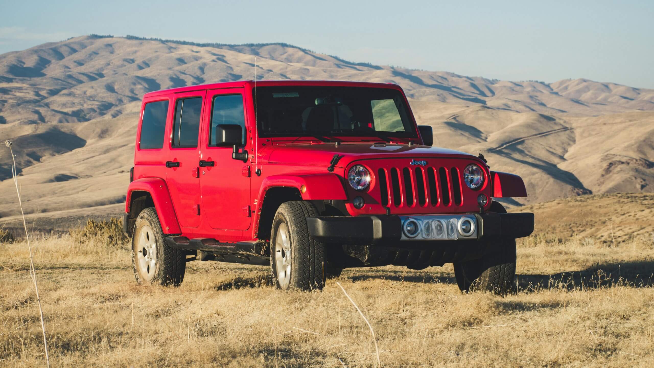Jeep Wrangler Merah diparkir di rumput kering dengan latar belakang perbukitan di bawah langit cerah