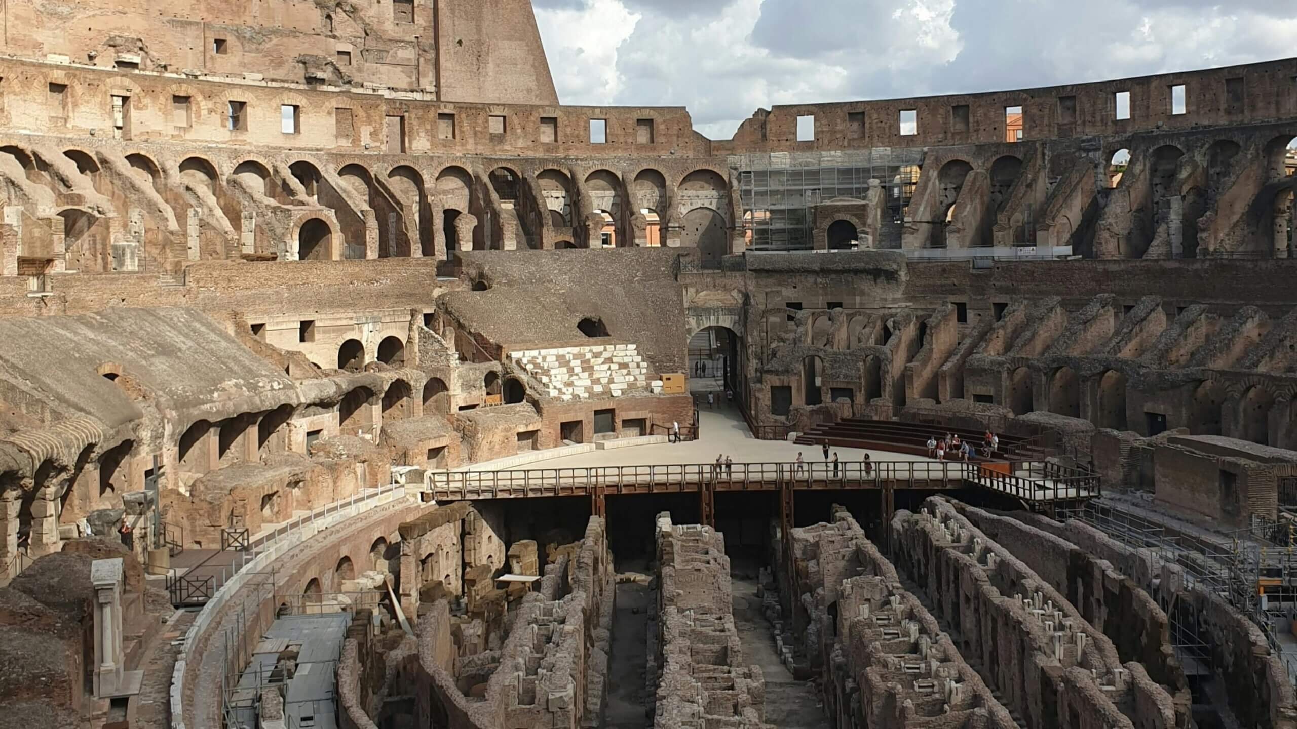 Pemandangan interior Colosseum Romawi kuno dengan struktur bawah tanah yang terlihat dan wisatawan berjalan di platform tinggi.