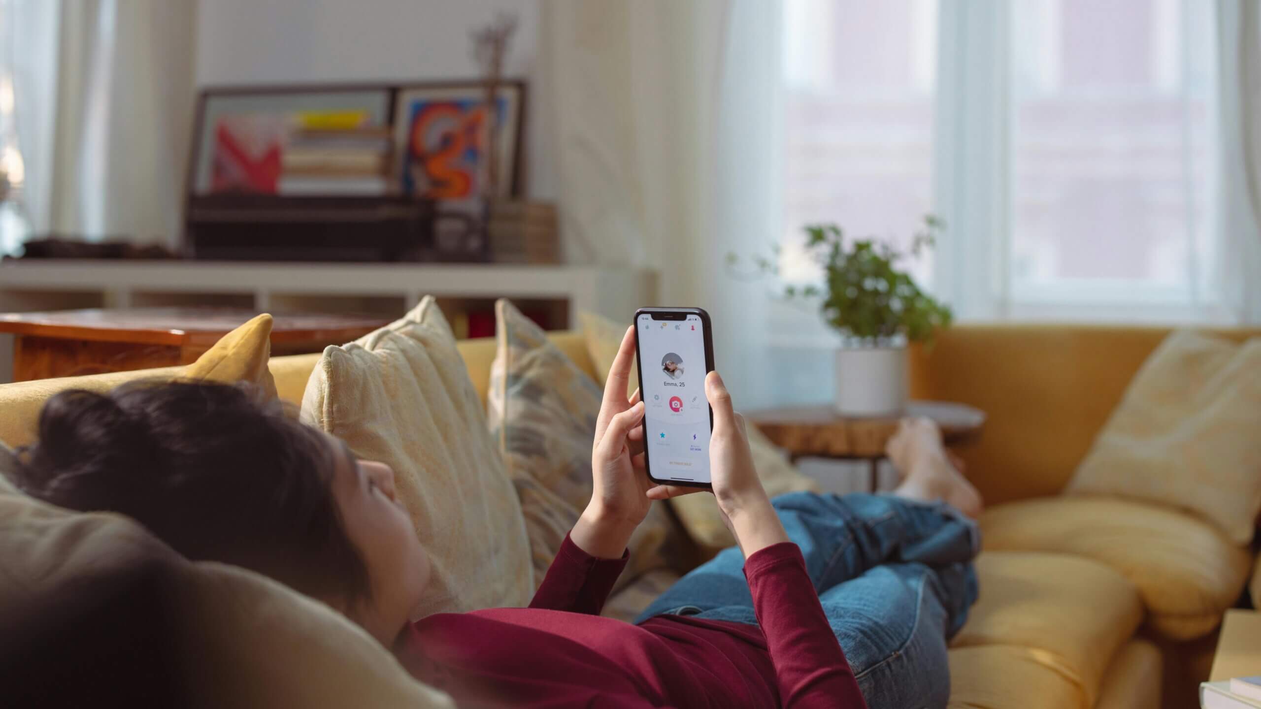 Seseorang berbaring di sofa kuning menggunakan smartphone yang menunjukkan profil aplikasi kencan Emma, 25.