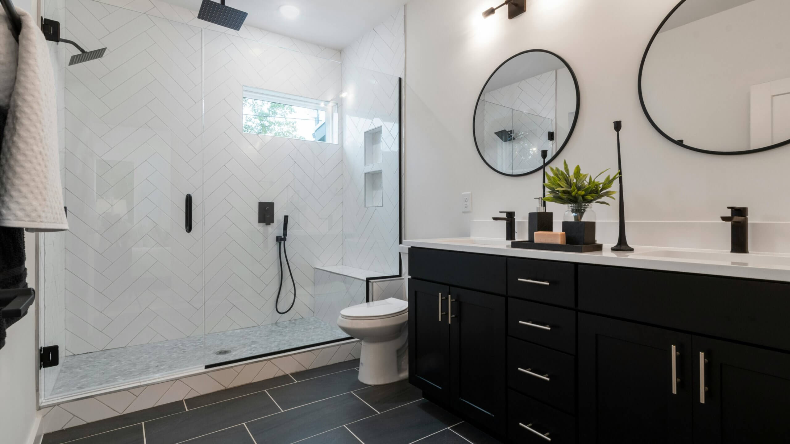 Kamar mandi modern dengan lemari hitam, cermin bundar ganda, pancuran kaca dengan ubin herringbone putih, dan perlengkapan hitam.