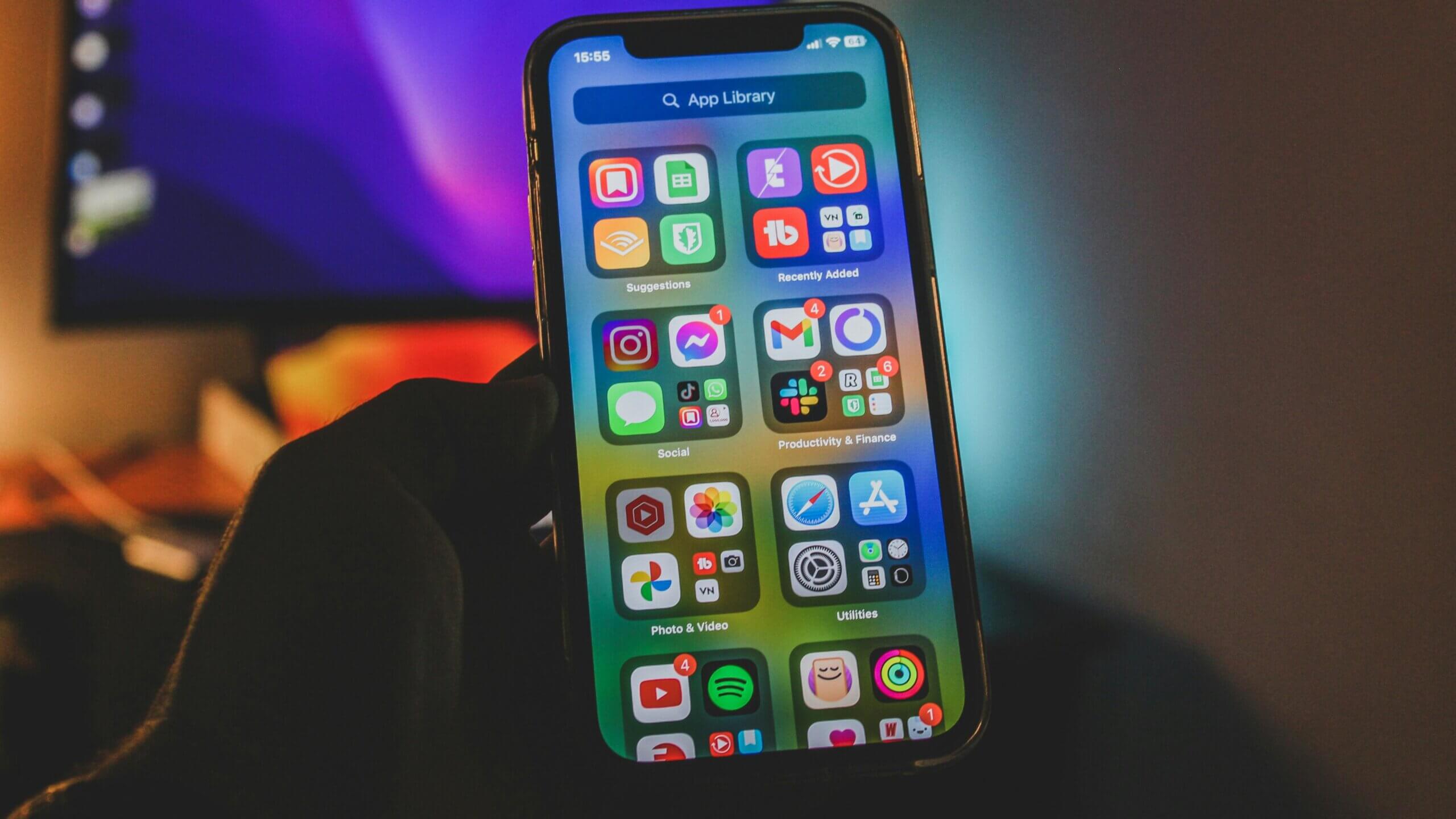 iPhone yang memegang tangan menampilkan Perpustakaan Aplikasi dengan folder aplikasi sosial, produktivitas, foto, dan utilitas terlihat