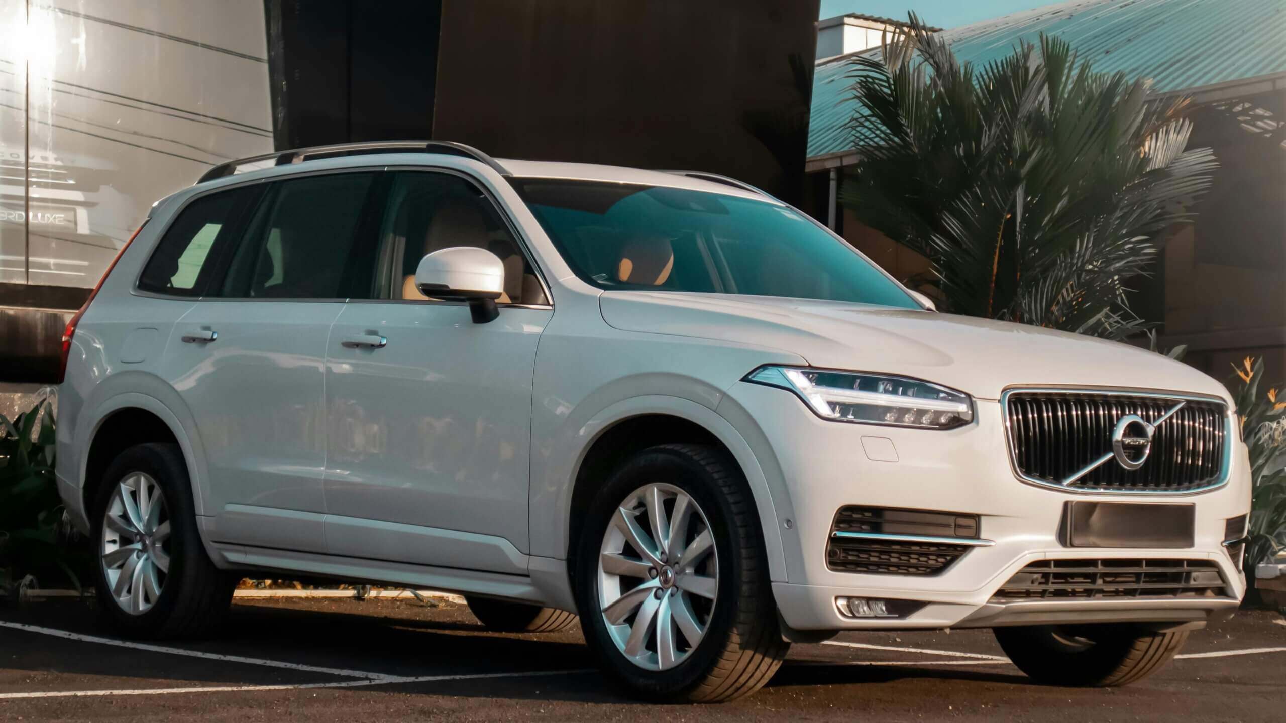 SUV Volvo XC90 putih diparkir di luar ruangan dekat tanaman dan bangunan di siang hari