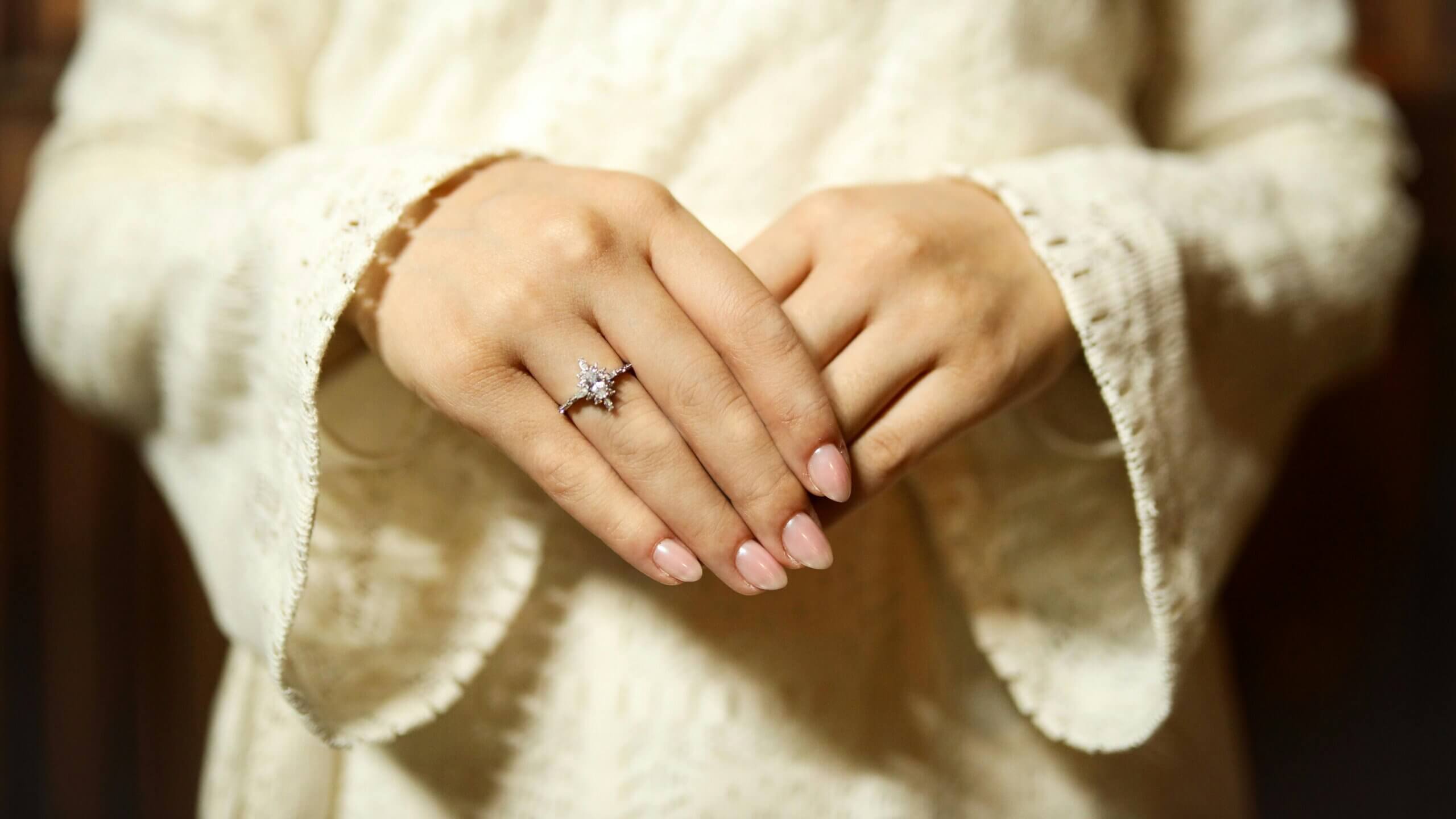 Tangan dengan cincin pertunangan berlian di jari manis kiri, mengenakan sweter rajutan berwarna krem.