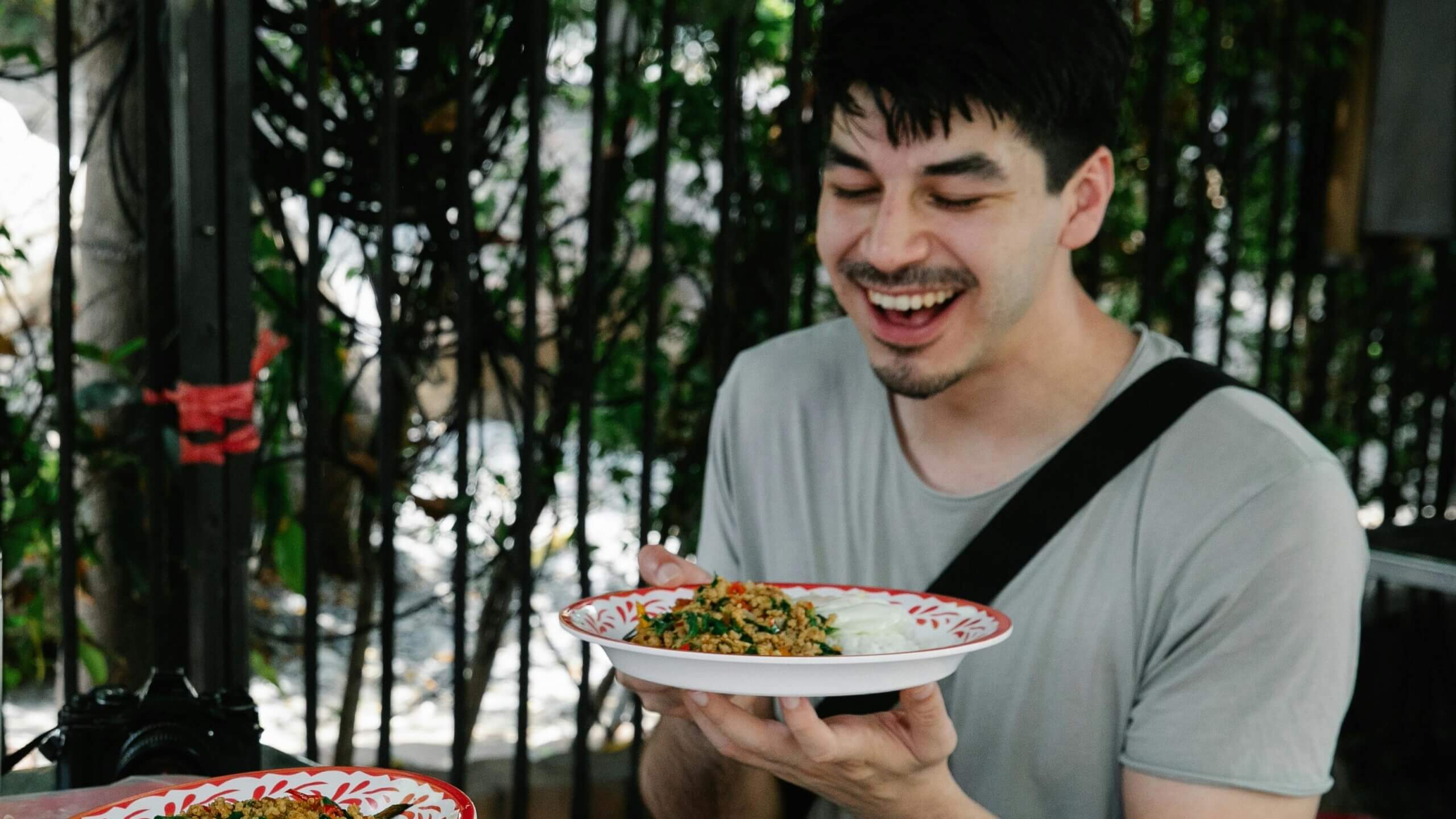 Pria tersenyum sambil memegang sepiring makanan dengan nasi dan sayur-sayuran di luar ruangan dekat pagar.