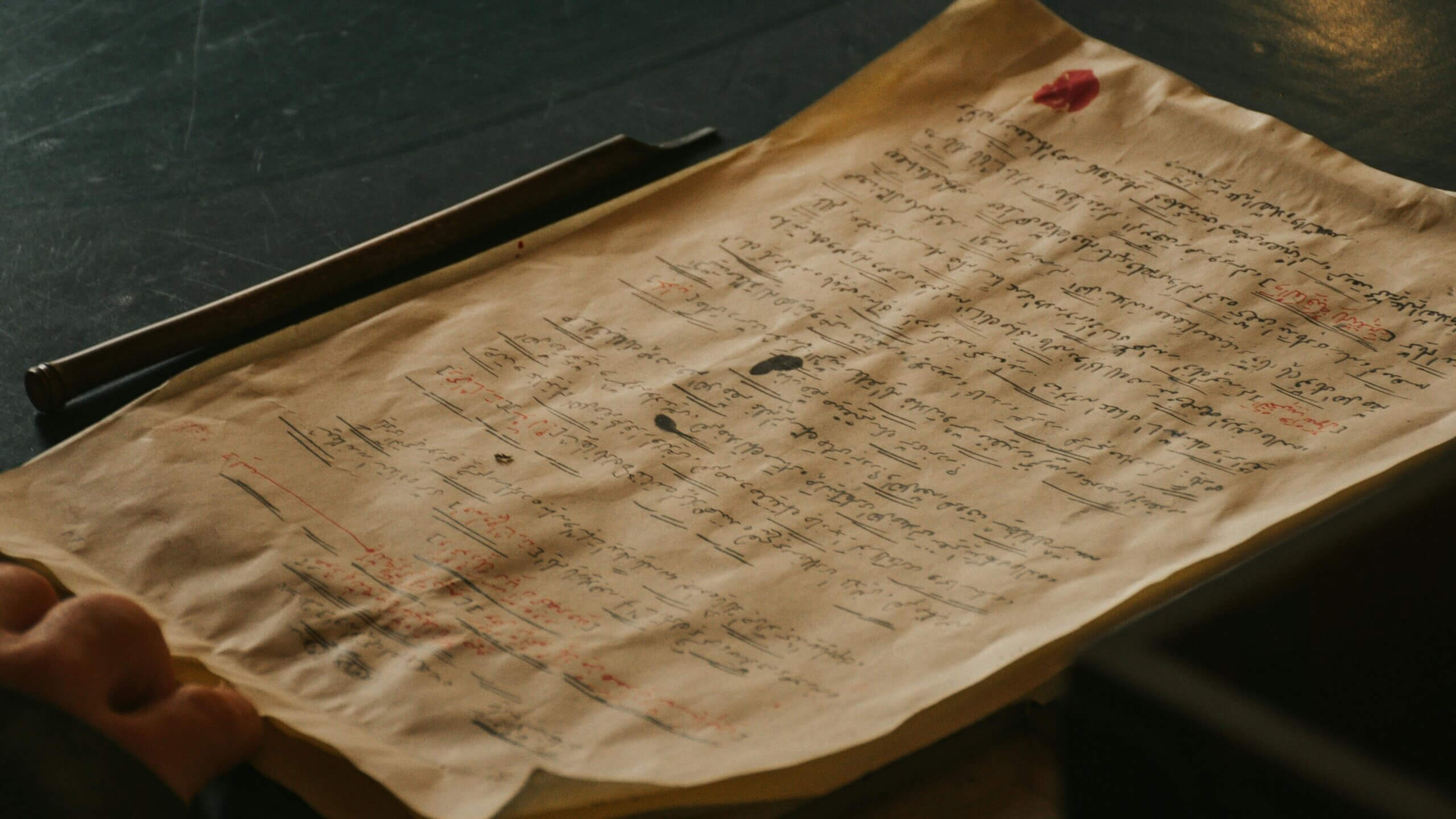 Dokumen tulisan tangan dengan tanda tinta merah dan hitam pada kertas tua di samping tongkat kayu pada permukaan gelap.