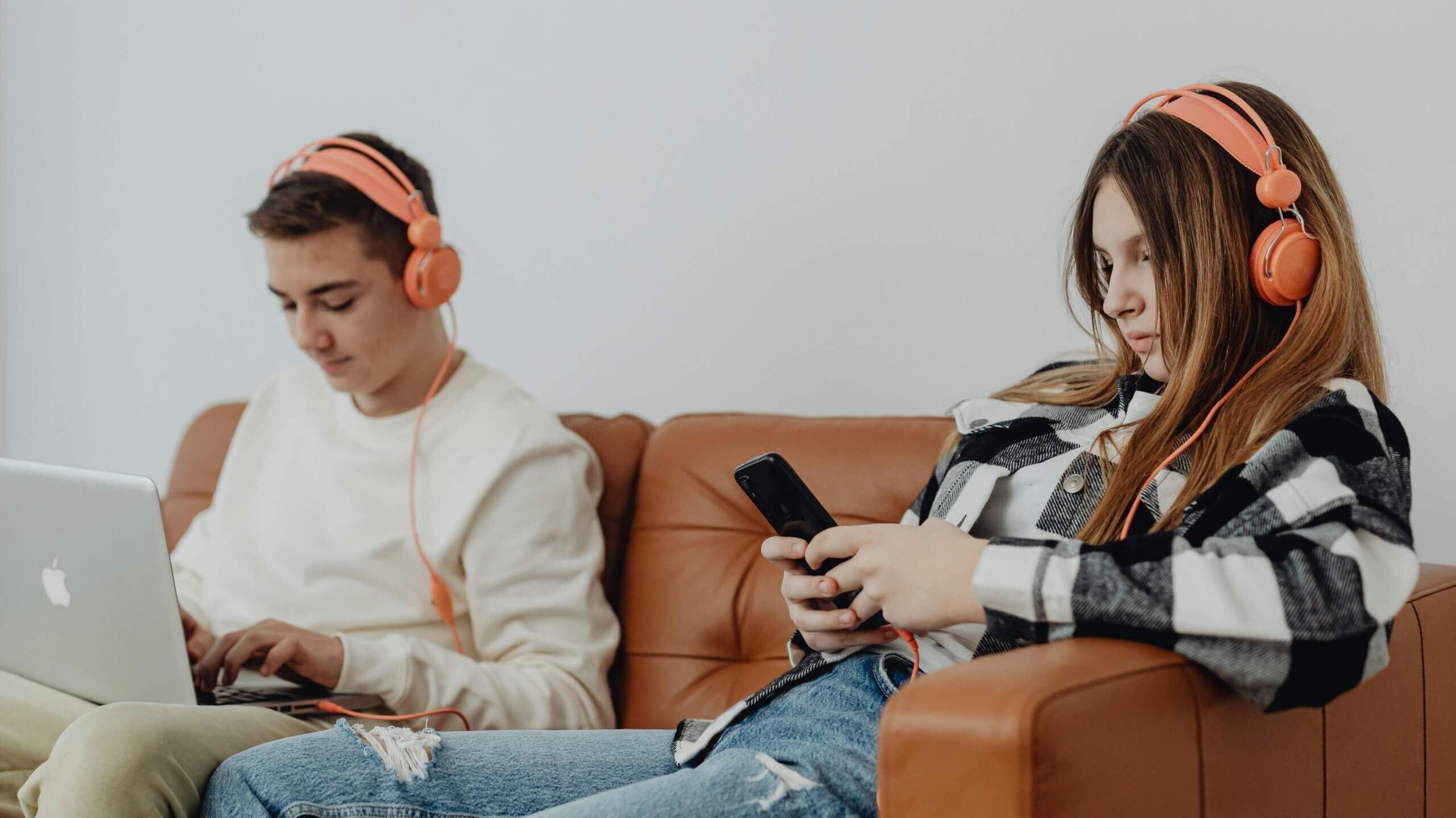 Remaja laki-laki menggunakan MacBook dan remaja perempuan berkemeja kotak-kotak menggunakan smartphone, keduanya mengenakan headphone oranye di sofa coklat.