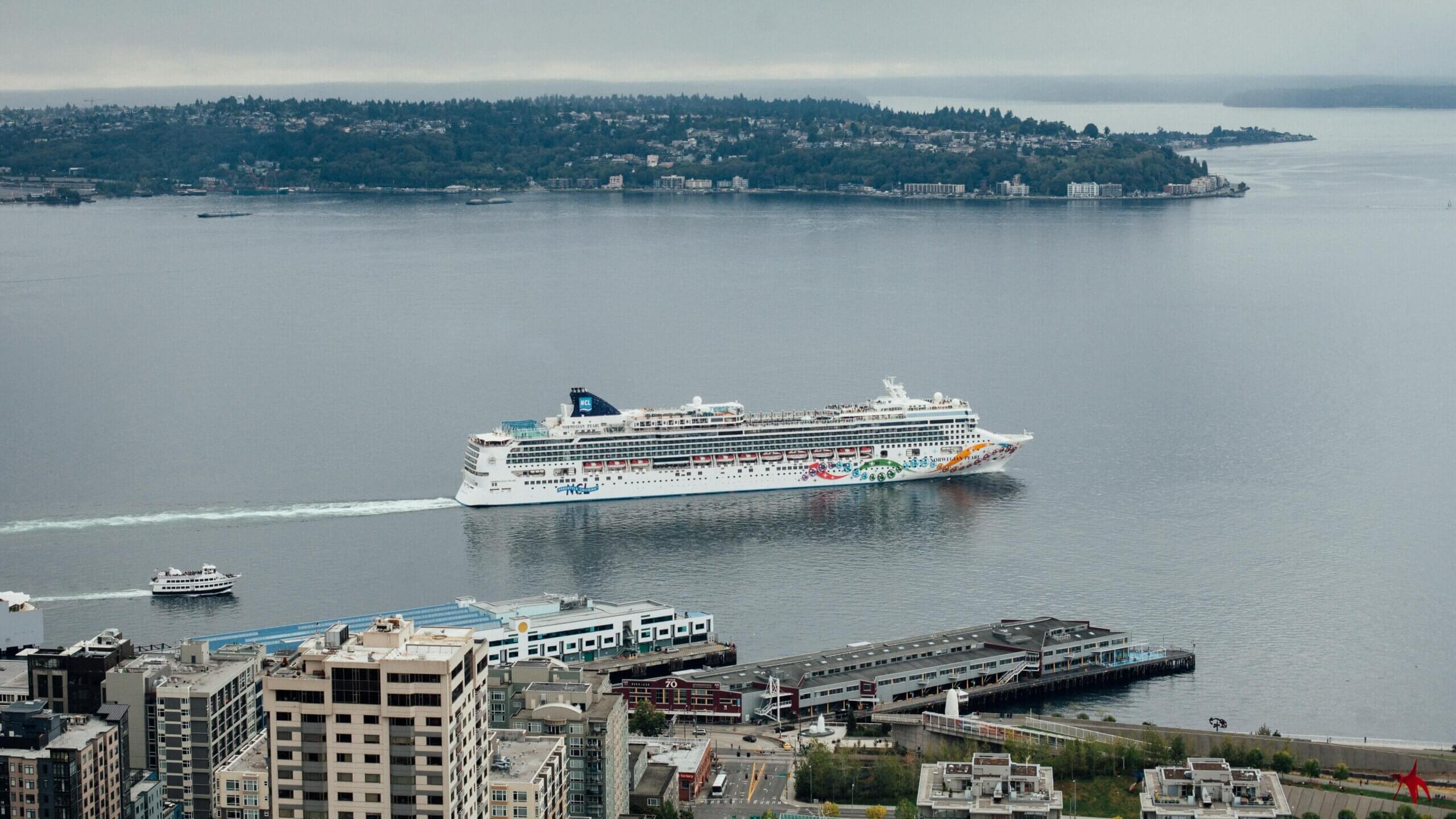 Kapal pesiar Mutiara Norwegia berlayar di dekat tepi laut kota dengan perahu kecil di dekatnya dan pulau berhutan di latar belakang.