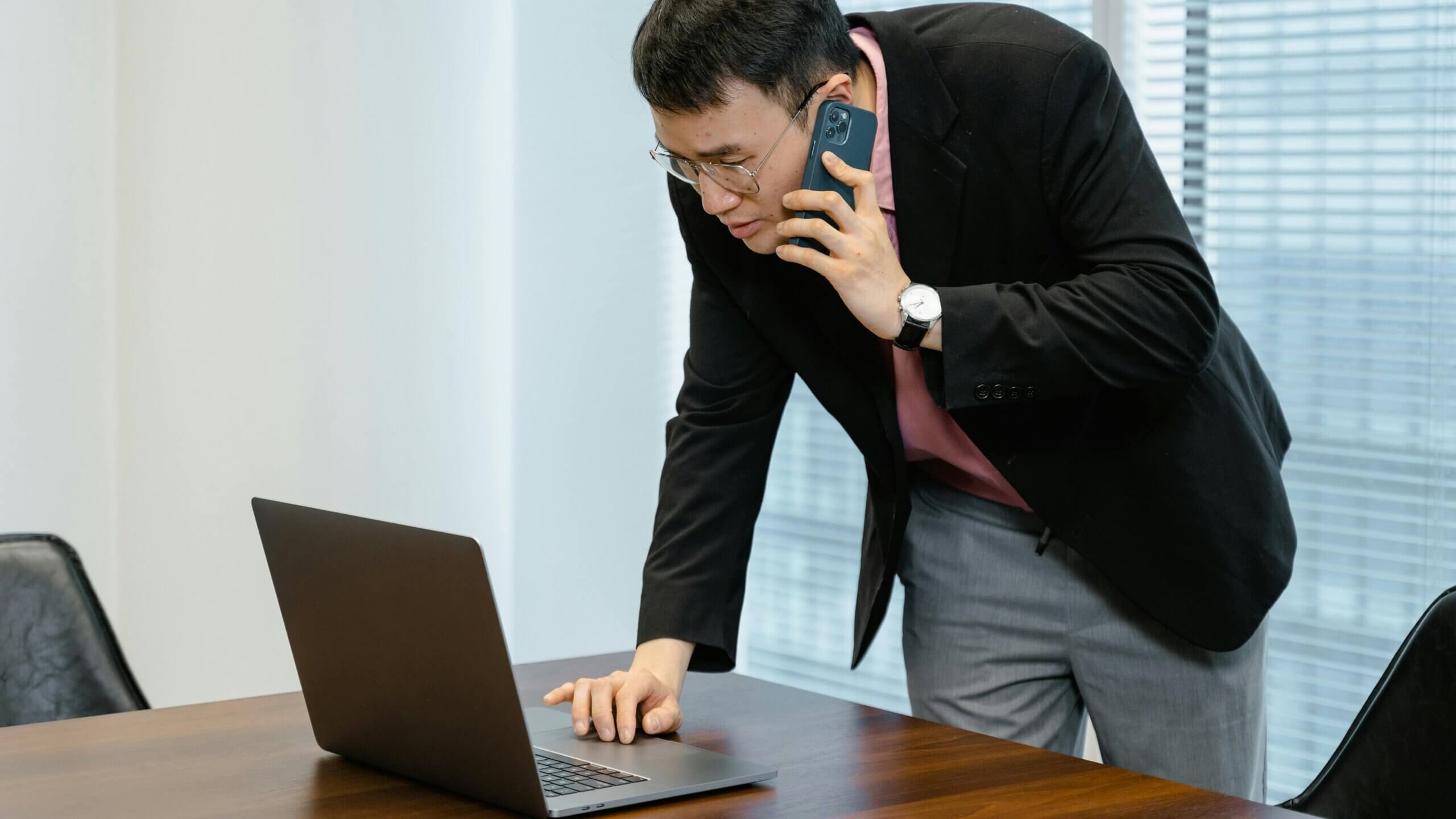 Pria berkacamata dan blazer hitam berbicara di smartphone sambil menggunakan laptop di meja kayu di kantor