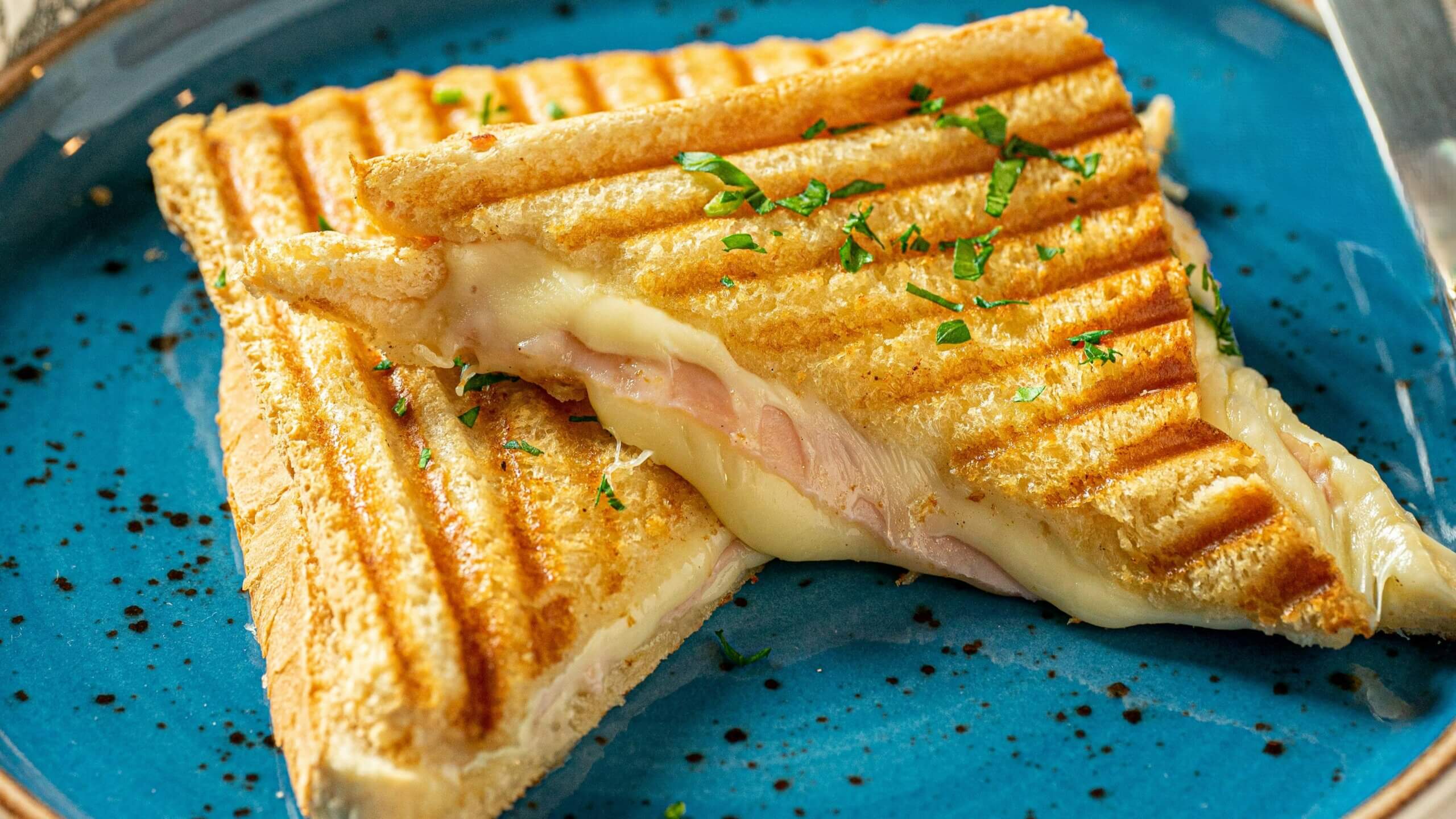 Bagian sandwich ham panggang dan keju leleh dihias dengan bumbu cincang di piring biru