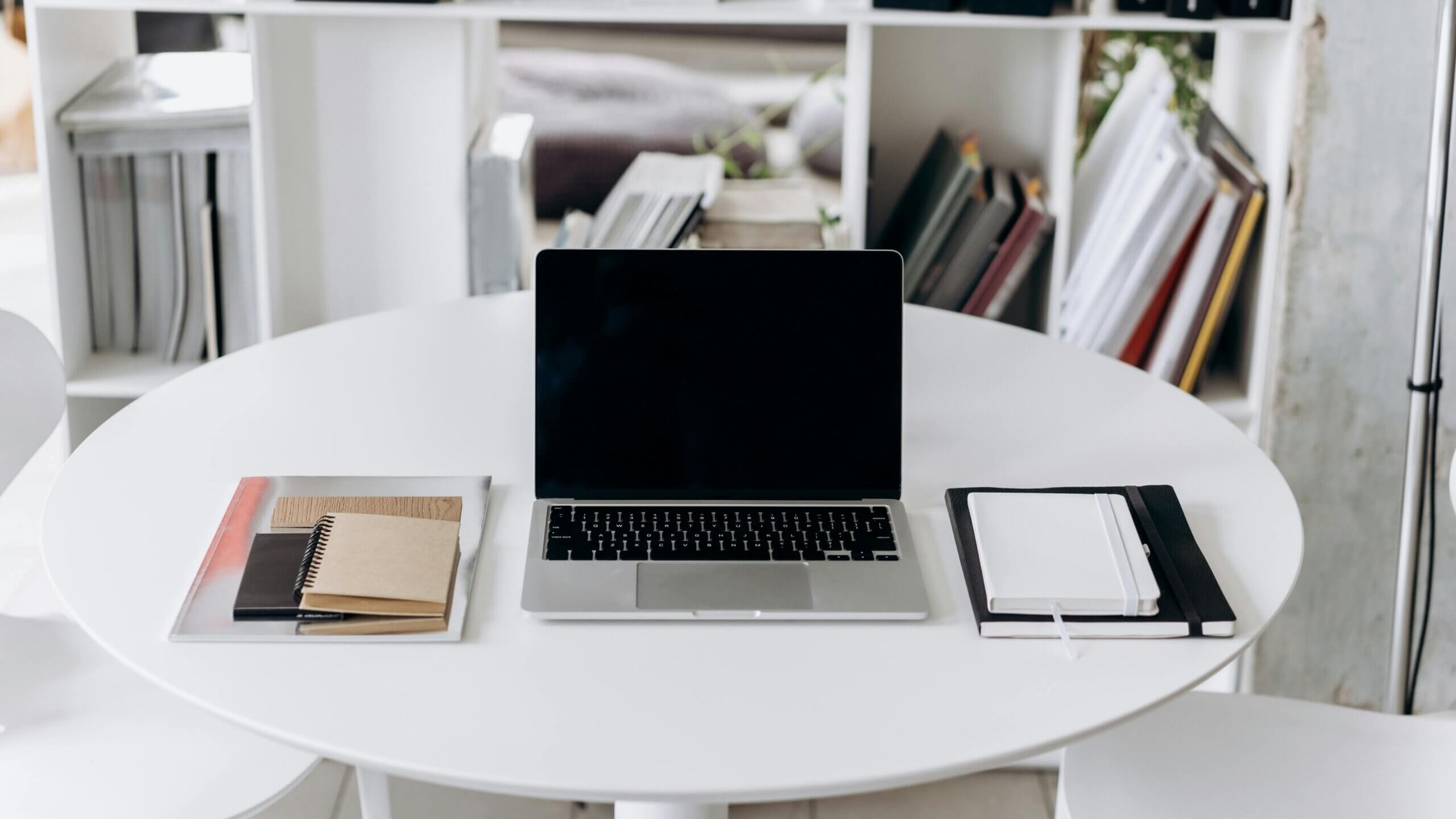 Laptop MacBook dengan layar hitam di atas meja bundar putih diapit oleh buku catatan dan folder dalam suasana kantor modern
