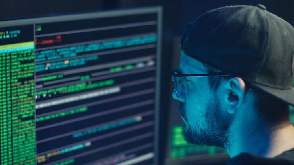 Pria berkacamata dan bertopi terbalik fokus pada layar komputer yang menampilkan kode dan data.