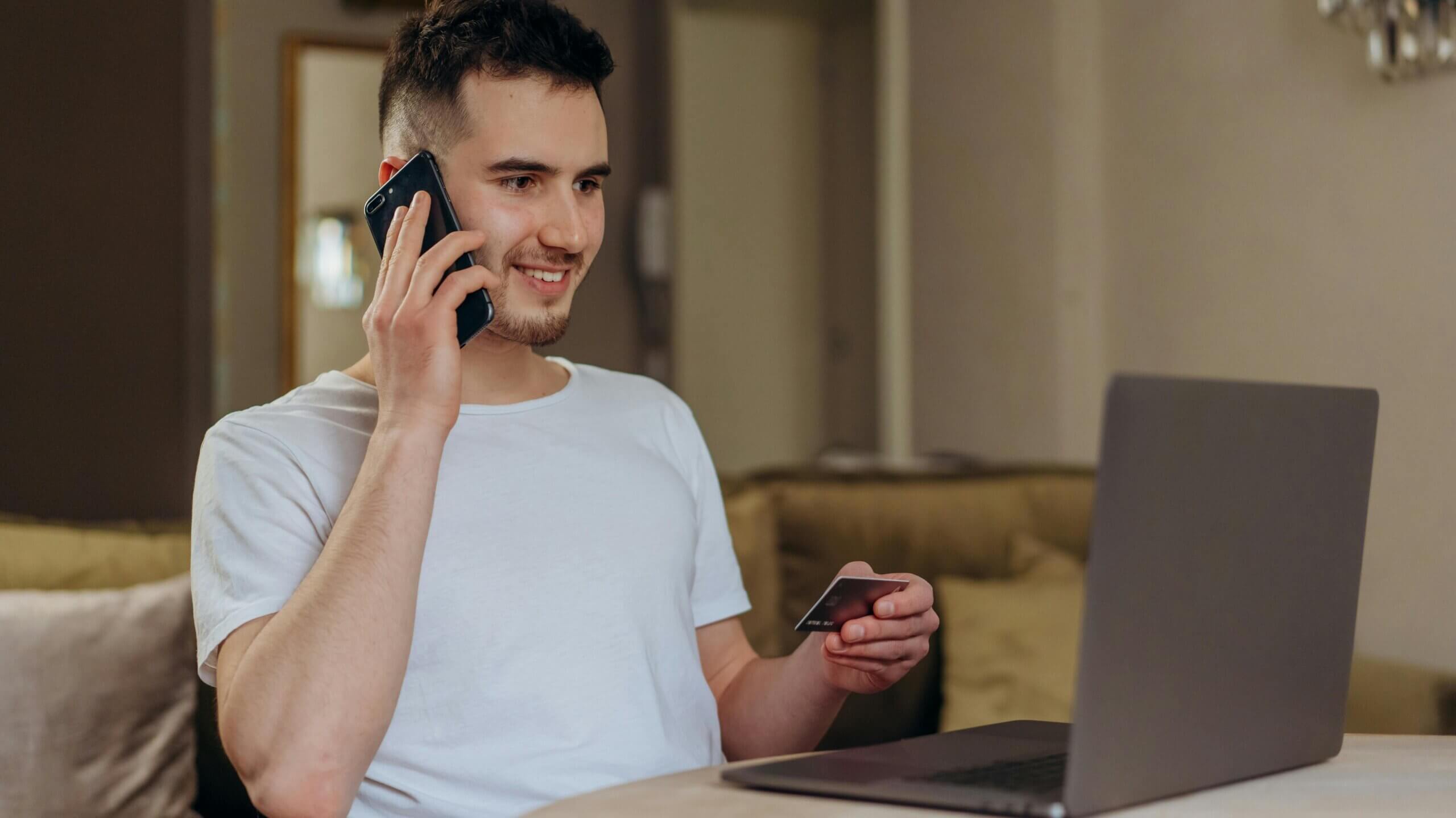 Pemuda berbaju putih memegang kartu kredit dan berbicara di telepon sambil menggunakan laptop di rumah