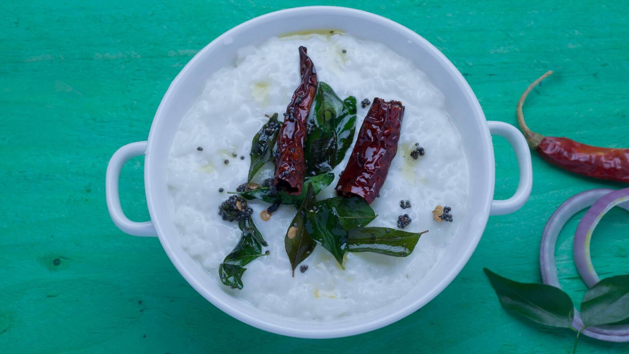 Nasi dadih yang dihias dengan cabai merah kering, daun kari, dan biji sawi dalam mangkuk putih dengan permukaan hijau.