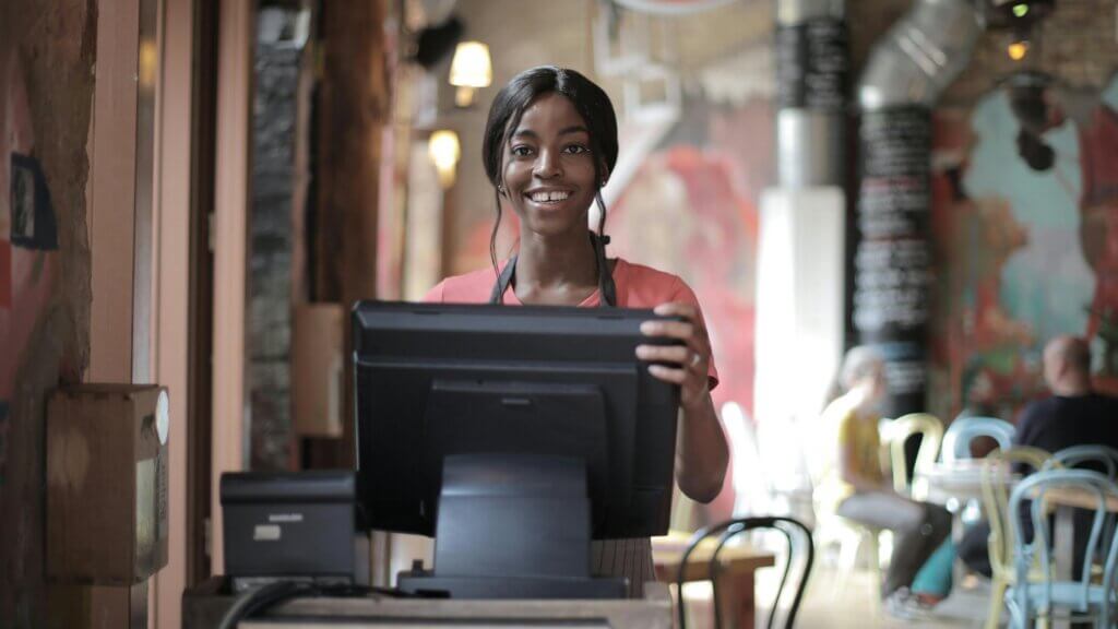 Wanita tersenyum mengenakan celemek berdiri di belakang mesin kasir di kafe dengan latar belakang pelanggan