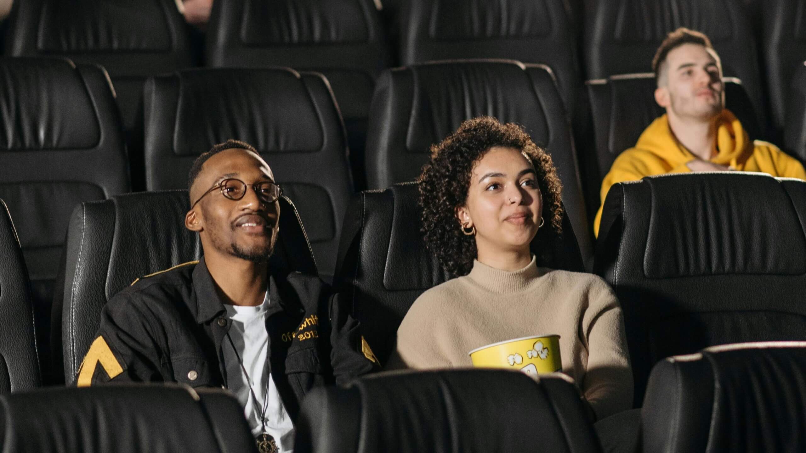 Tiga orang duduk di bioskop, salah satunya memegang ember popcorn kuning.