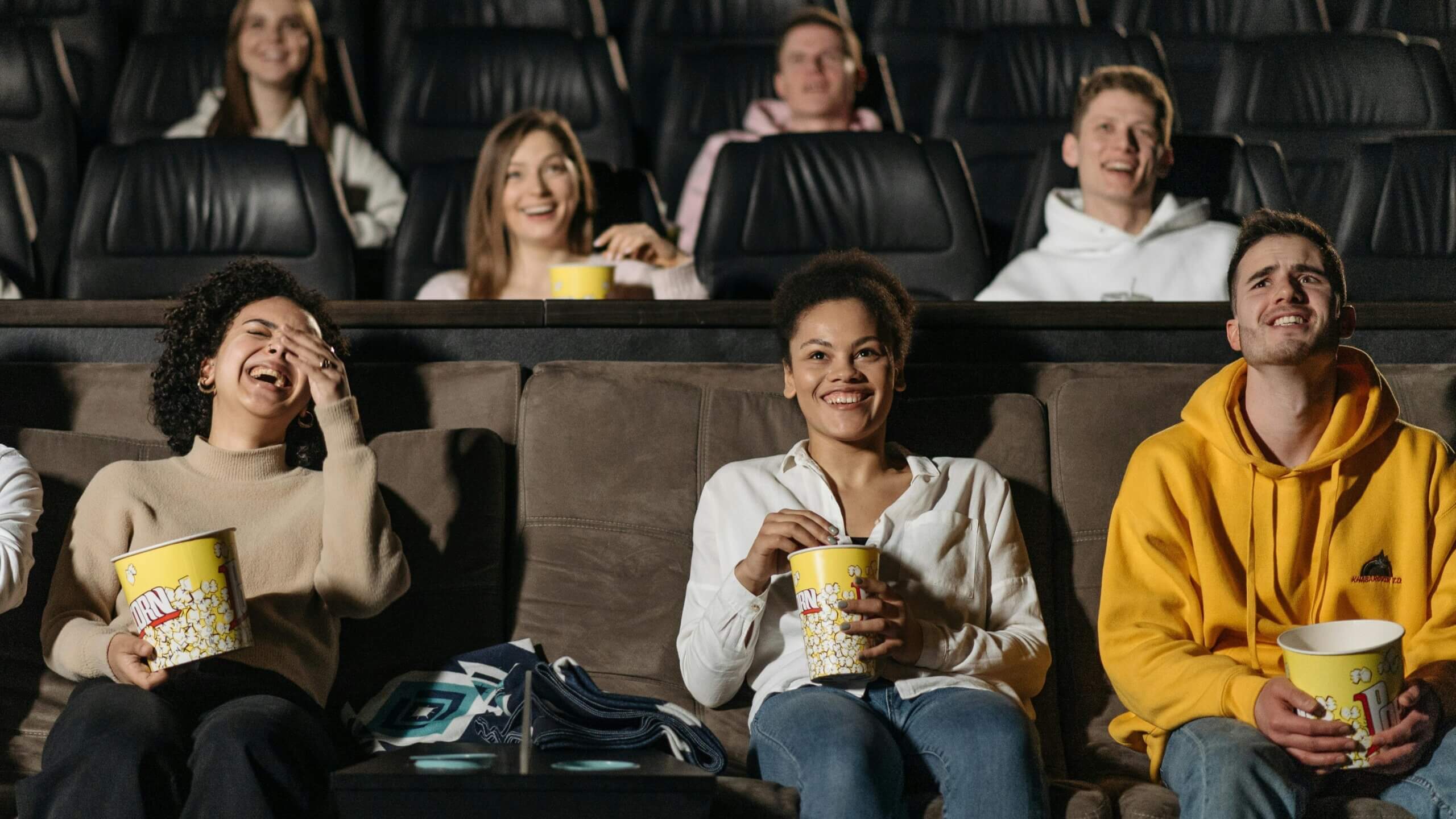 Sekelompok anak muda tertawa dan makan popcorn di bioskop dengan tempat duduk berjenjang