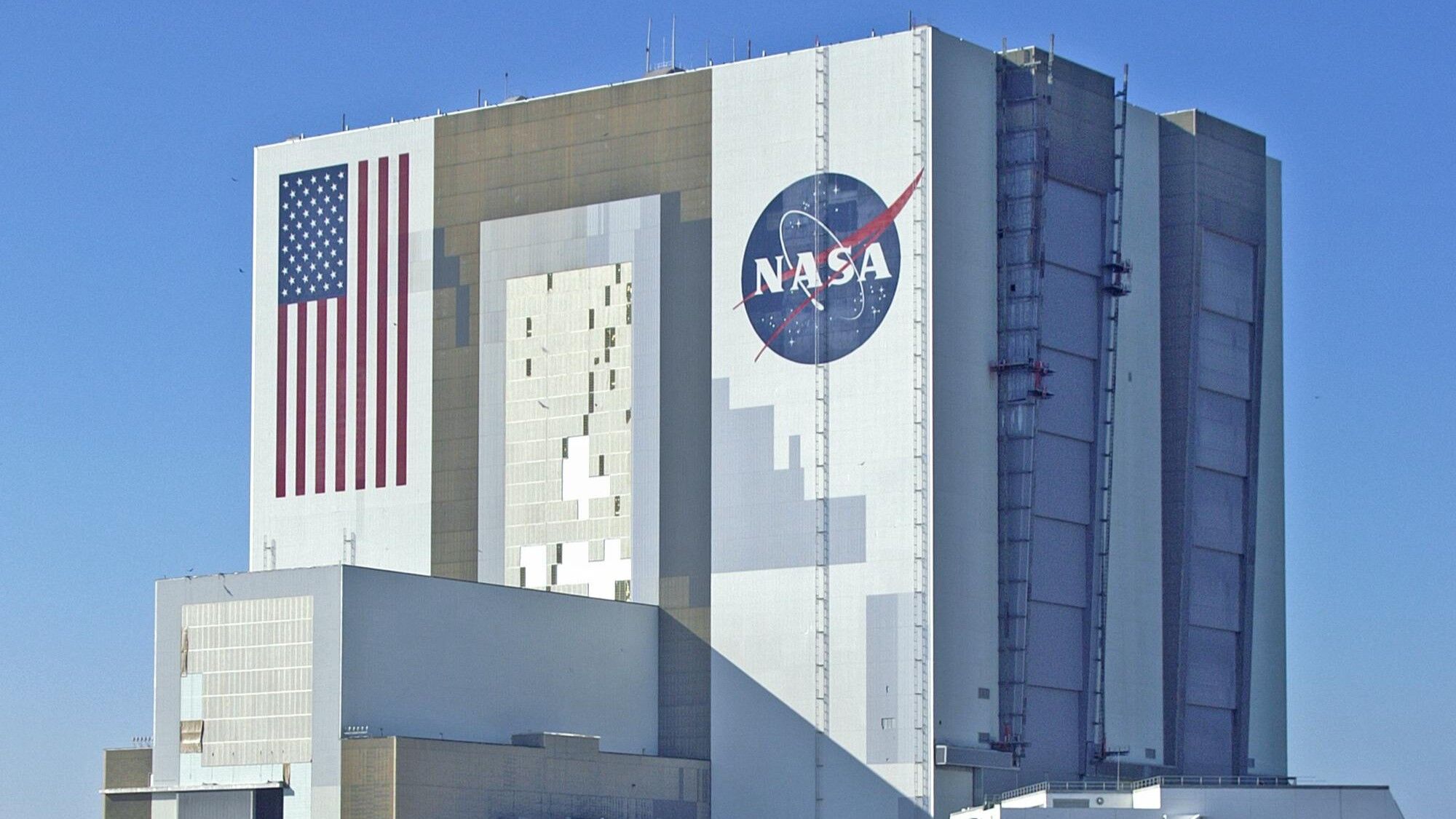 Gedung Perakitan Kendaraan NASA dengan bendera Amerika besar dan logo NASA di bagian luarnya di bawah langit biru cerah