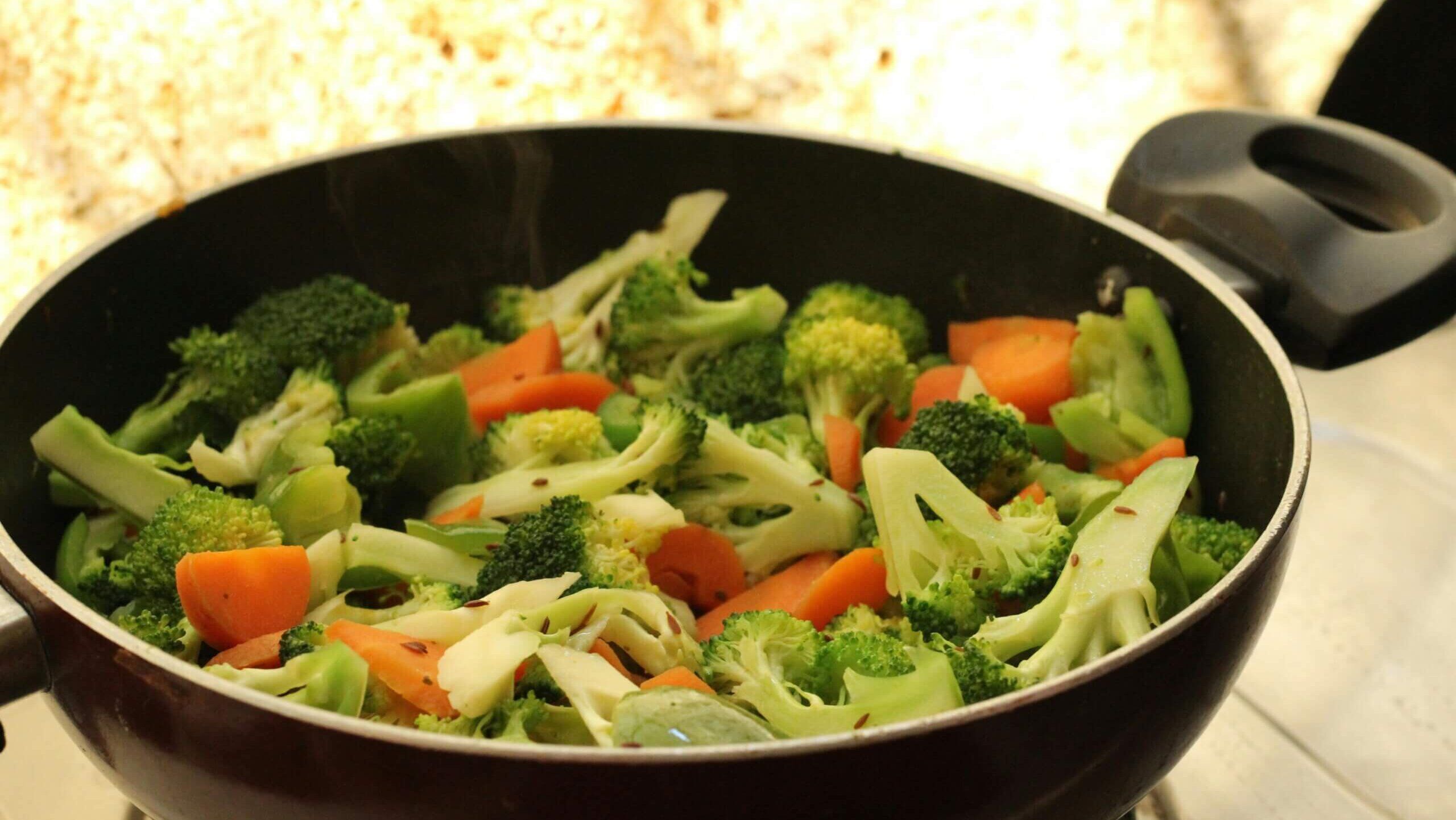 Mengukus campuran sayuran termasuk brokoli, wortel, dan paprika hijau yang dimasak dalam wajan hitam.