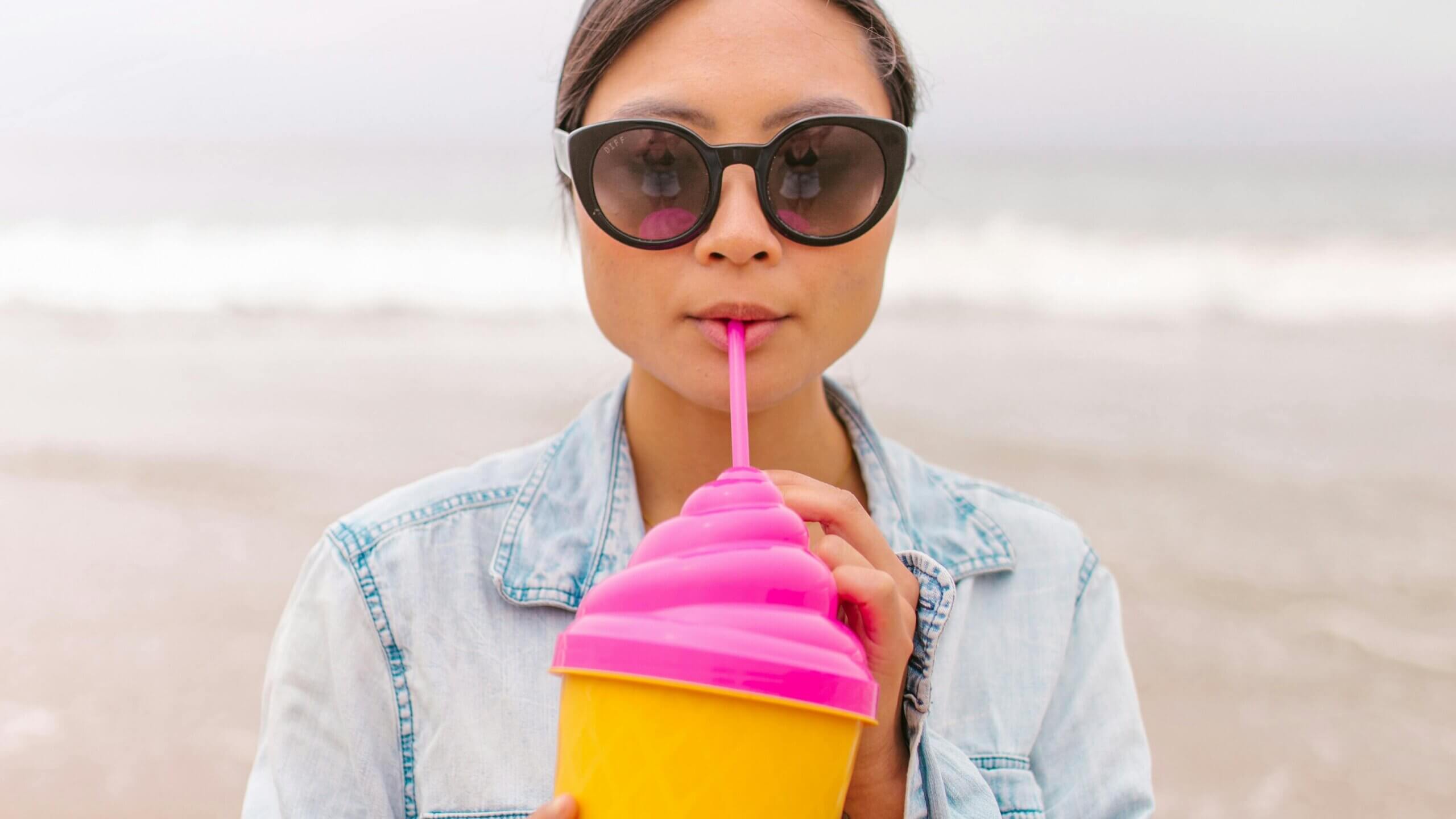 Wanita mengenakan kacamata hitam dan jaket denim minum dari cangkir merah muda dan kuning dengan sedotan di pantai