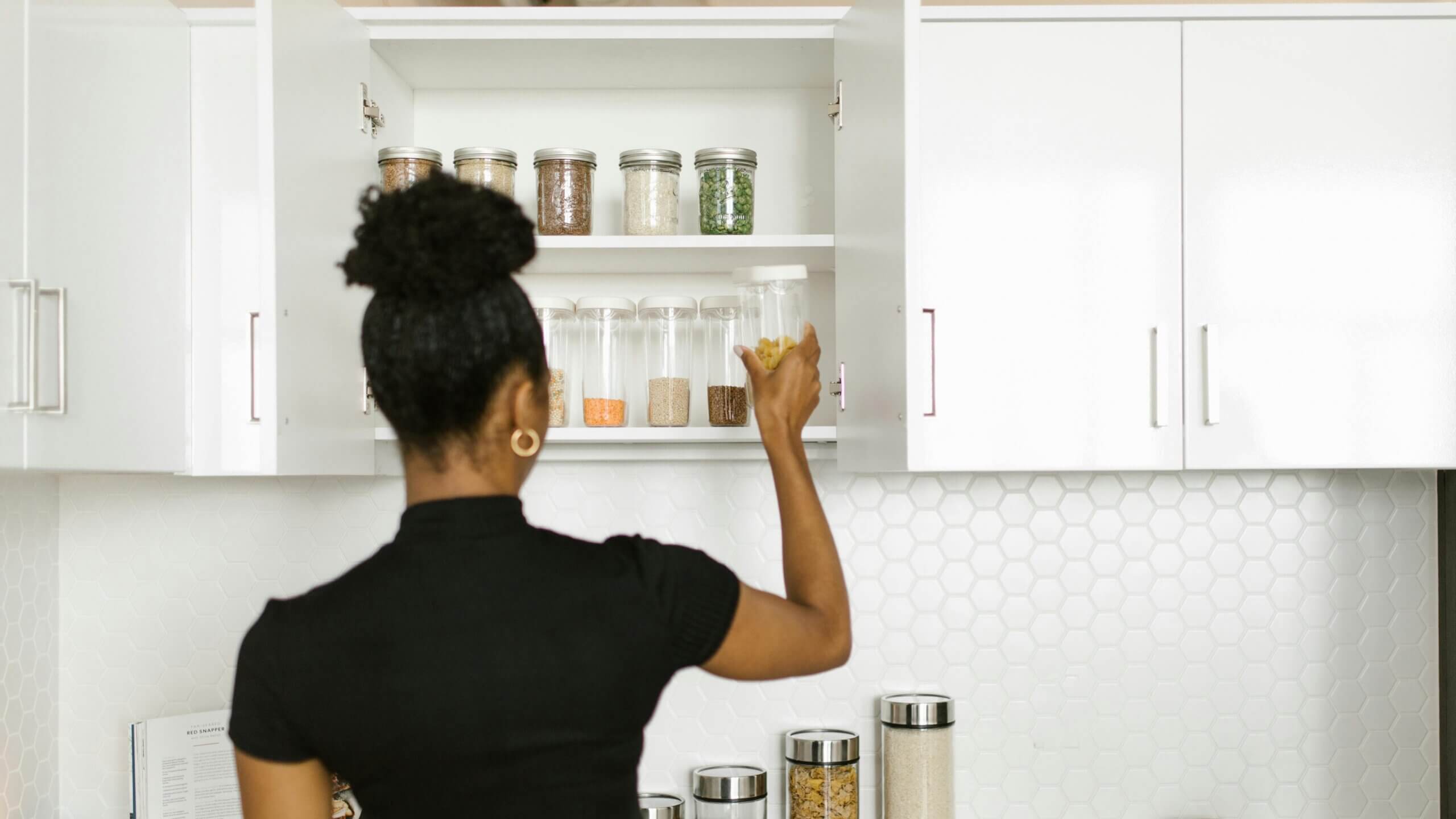 Wanita berkemeja hitam meraih sebotol pasta dari lemari dapur putih dengan wadah terorganisir.