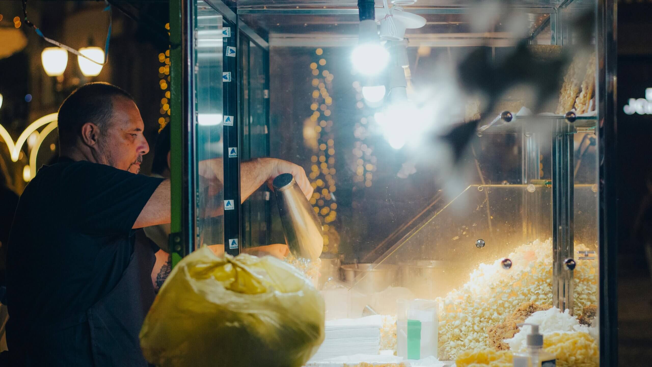 Seorang pria menyendok popcorn dari mesin popcorn di malam hari dengan lampu senar di latar belakang