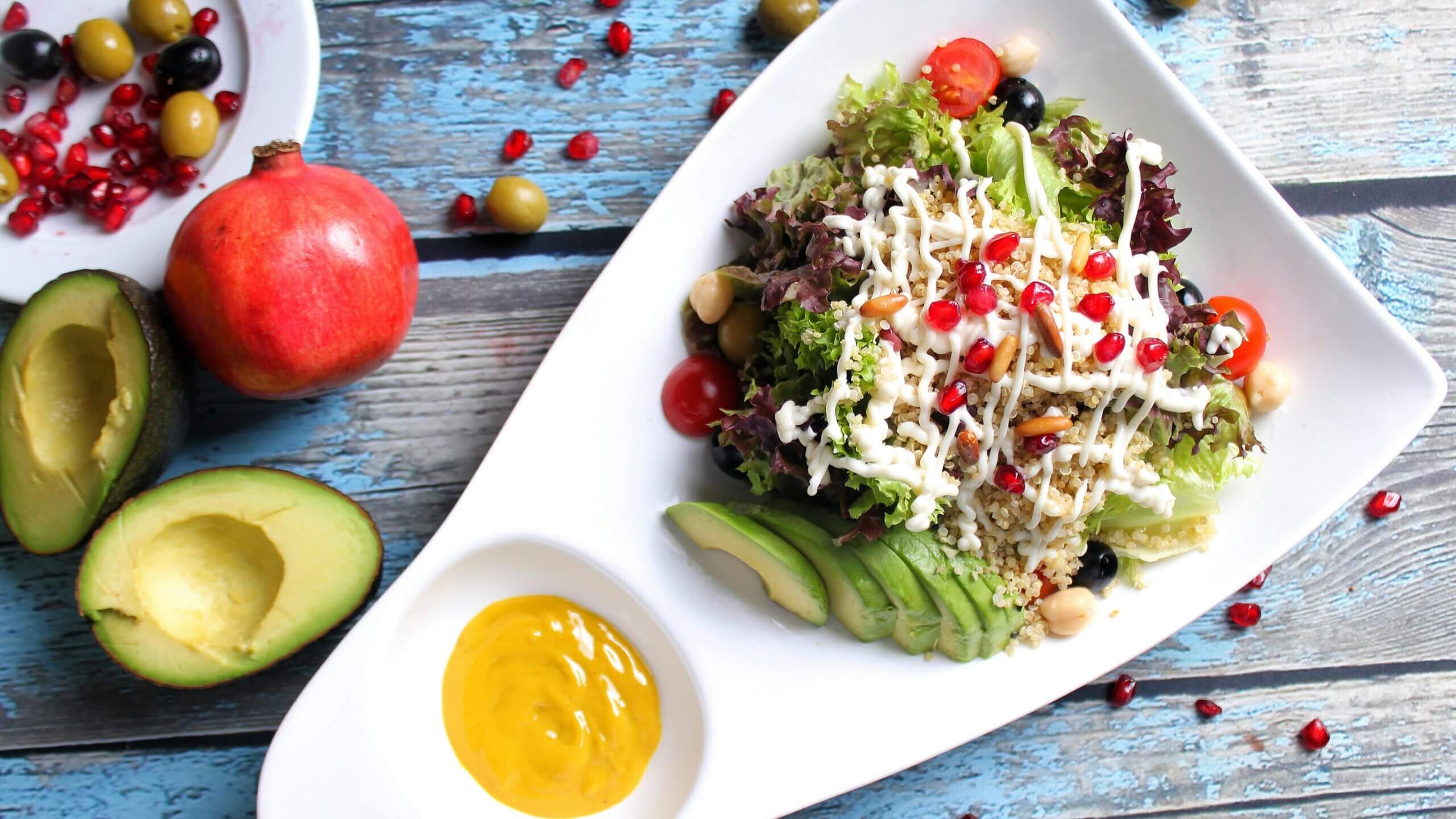 Salad Quinoa dengan selada, tomat ceri, zaitun, biji delima, irisan alpukat, dan saus mustard di piring putih