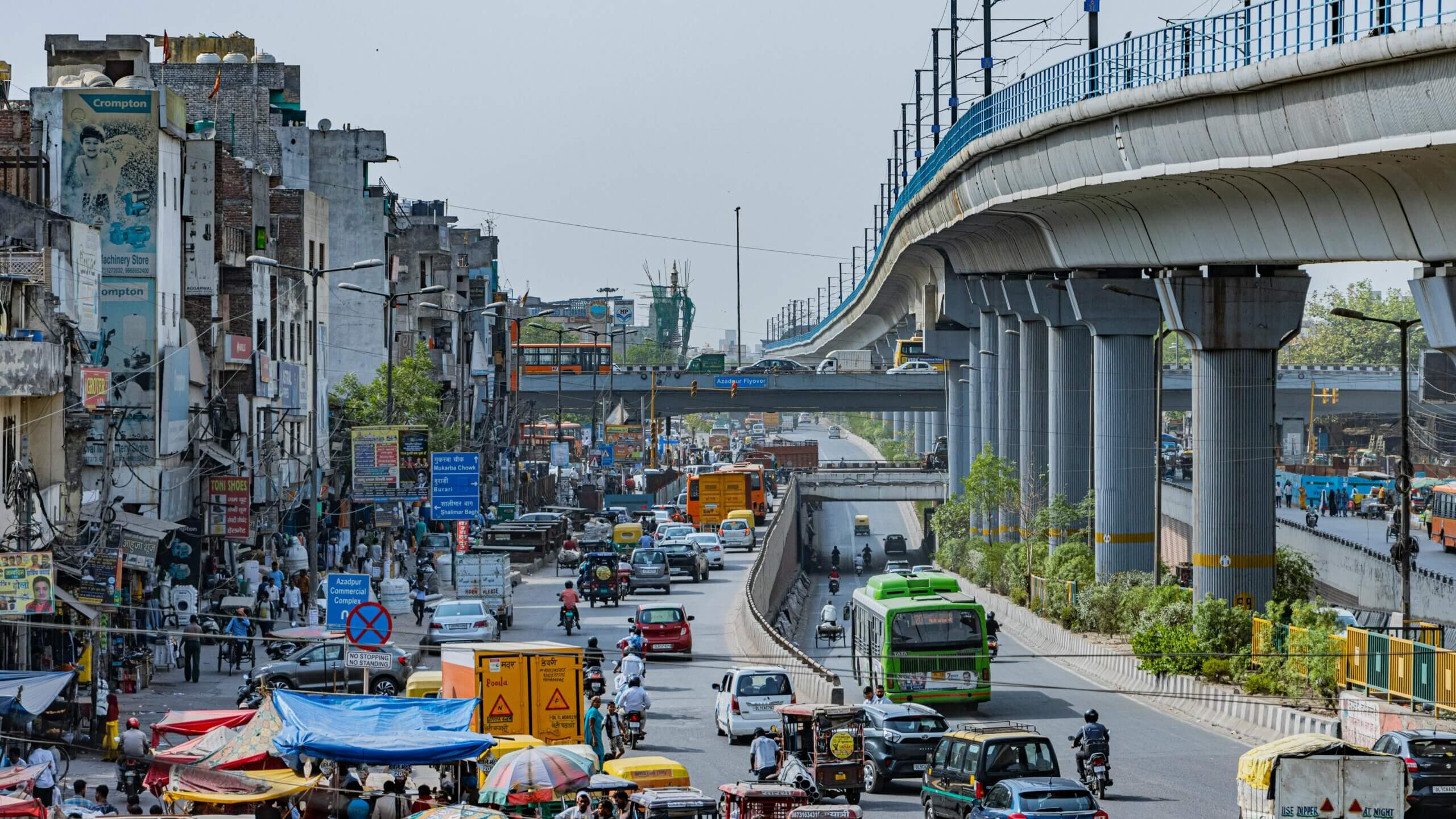 Jalan perkotaan yang sibuk dengan lalu lintas padat, sepeda motor, bus, dan pejalan kaki di bawah jalur kereta metro dan jalan layang yang ditinggikan.