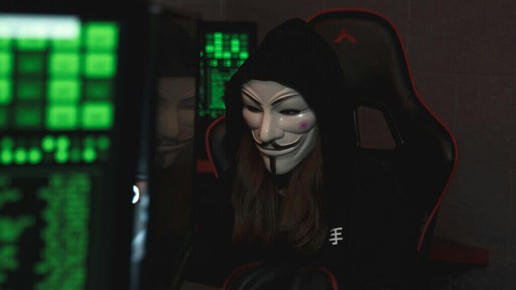 Orang yang memakai topeng Guy Fawkes dan hoodie hitam duduk di kursi permainan dengan kode hijau di layar di dekatnya