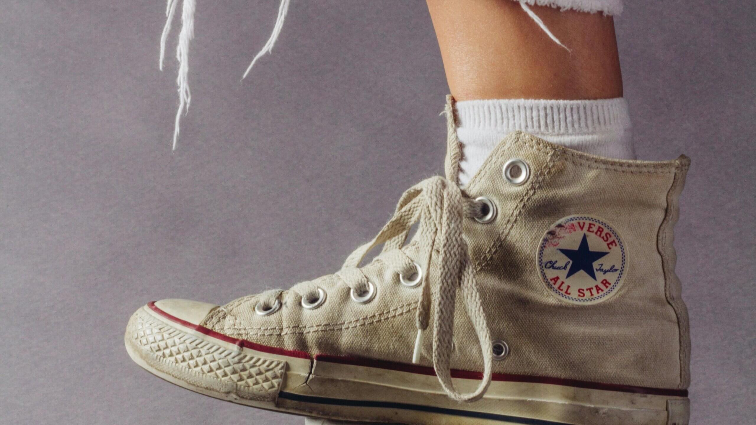 Sneaker Converse Chuck Taylor All Star berwarna krem dengan kaus kaki putih dan jeans berjumbai dengan latar belakang abu-abu