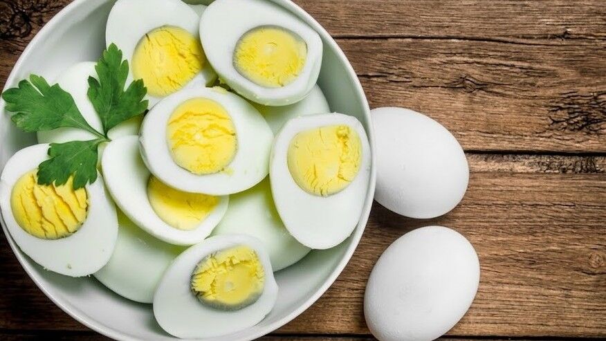 Semangkuk telur rebus yang dibelah dua dihias dengan peterseli di samping dua telur utuh di permukaan kayu