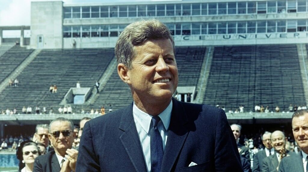 John F. Kennedy tersenyum dalam setelan jas di acara stadion dengan latar belakang penonton