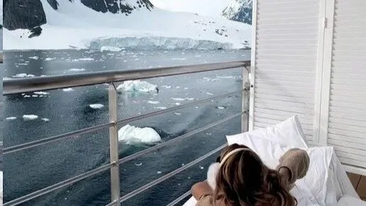 Orang berbaring di tempat tidur di balkon menghadap air es dan pegunungan yang tertutup salju.
