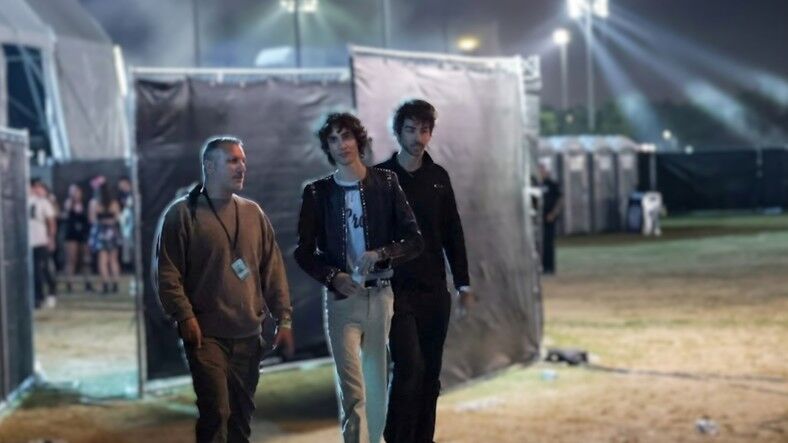 Tiga pria berjalan di luar ruangan pada malam hari dekat tenda acara dan toilet portabel