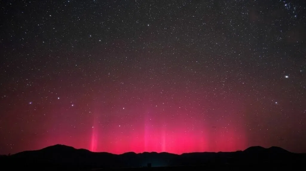 Aurora borealis merah bersinar di atas siluet gunung gelap di bawah langit malam berbintang.