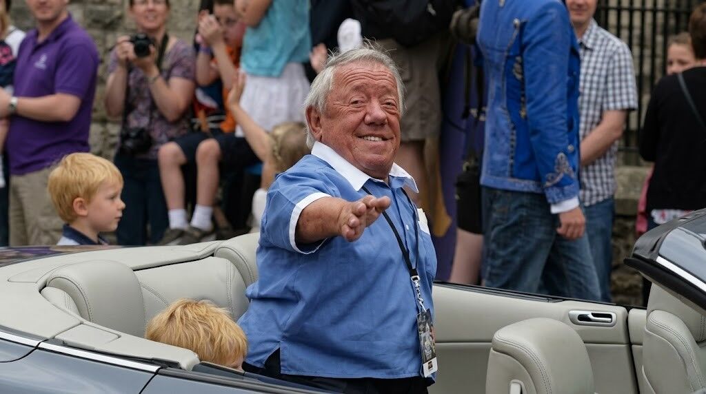 Pria tua berkemeja biru melambaikan tangan dari mobil konvertibel saat acara publik dengan penonton di latar belakang