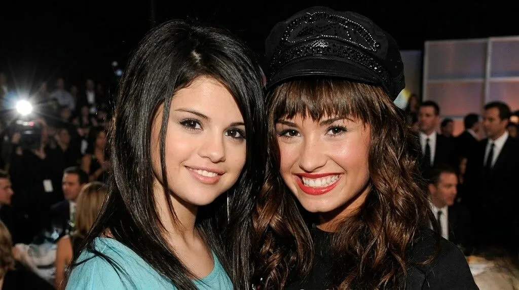 Selena Gomez dan Demi Lovato tersenyum bersama di sebuah acara formal dengan latar belakang orang-orang.