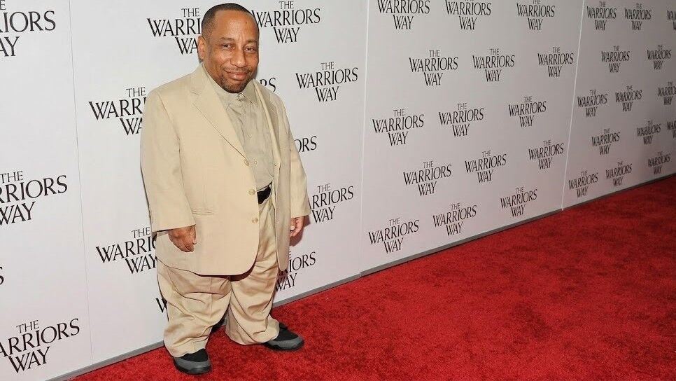 Pria berjas krem ​​berdiri di karpet merah dengan latar belakang acara The Warriors Way