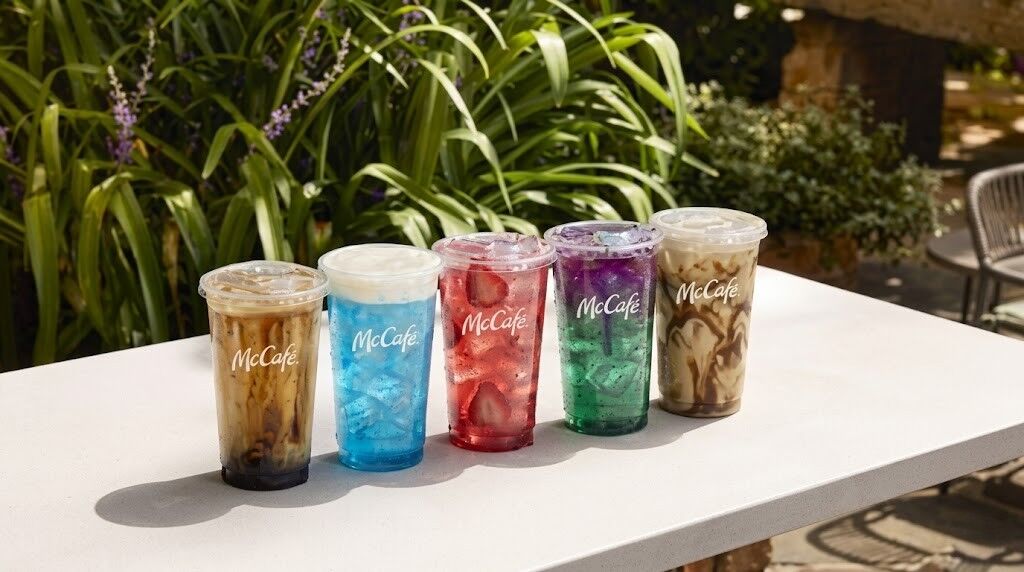 Lima minuman es McCafé dalam cangkir bening dengan lapisan berwarna-warni di atas meja putih di luar ruangan dengan tanaman hijau di latar belakang