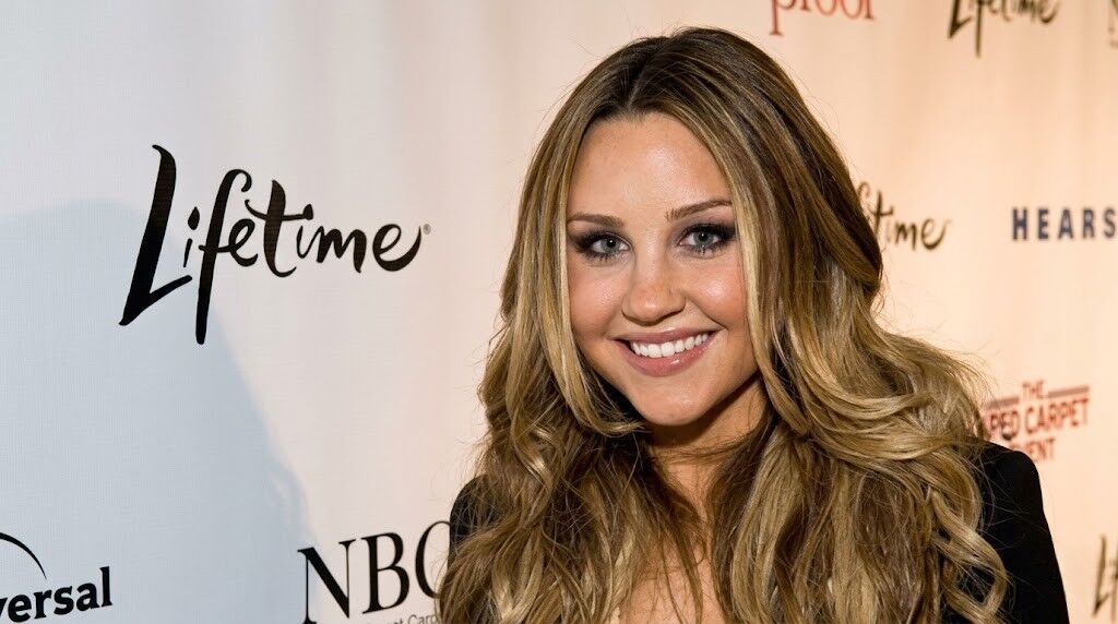 Amanda Bynes tersenyum di acara Seumur Hidup dengan logo NBCUniversal sebagai latar belakang