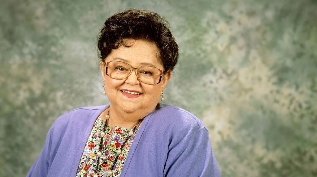 Wanita tua tersenyum berkacamata mengenakan kardigan ungu dan blus bunga dengan latar belakang hijau berbintik-bintik.