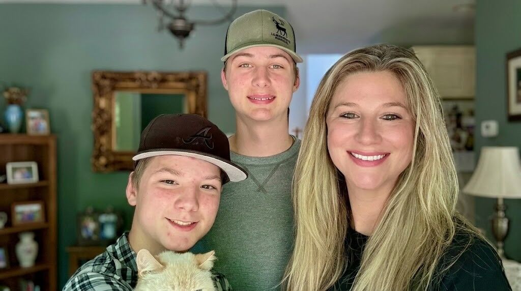 Tiga orang tersenyum, dua remaja laki-laki mengenakan topi baseball dan seorang wanita berambut pirang, dengan seekor kucing di lingkungan rumah.